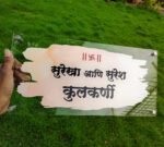 Hand holding a transparent Kulkarni Marathi watercolour name plate over green grass showing the clear acrylic