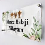 Angled close-up of transparent Shree Balaji Nilayam acrylic name plate with gold symbols and floral corner